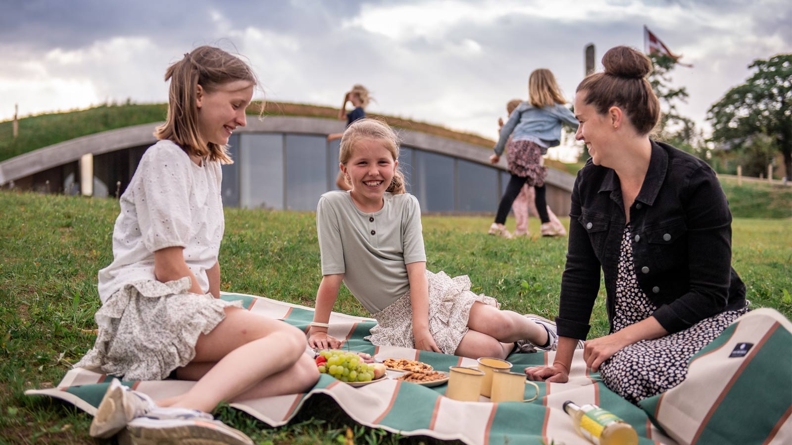 Eine Mutter und ihre beiden Töchter sitzen auf einer Decke und essen Snacks. Hinter ihnen befindet sich das Besucherzentrum, das in einen Erdwall gebaut ist.