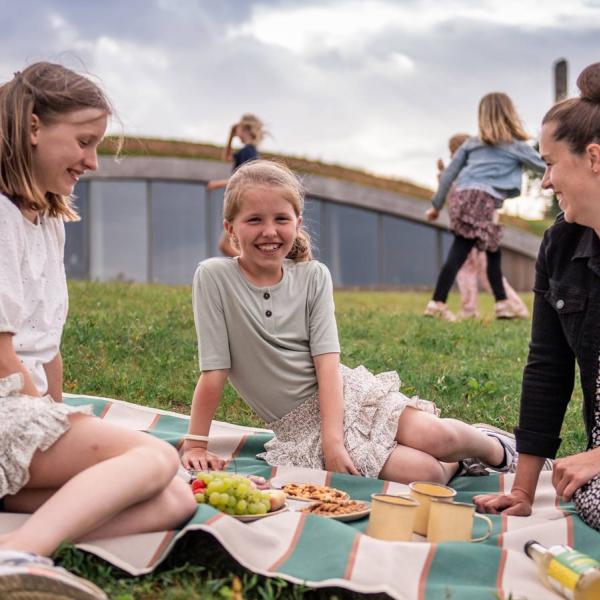 A mother and her two daughters sit on a blanket eating snacks. Behind them is the visitor centre, built into an earth bank.