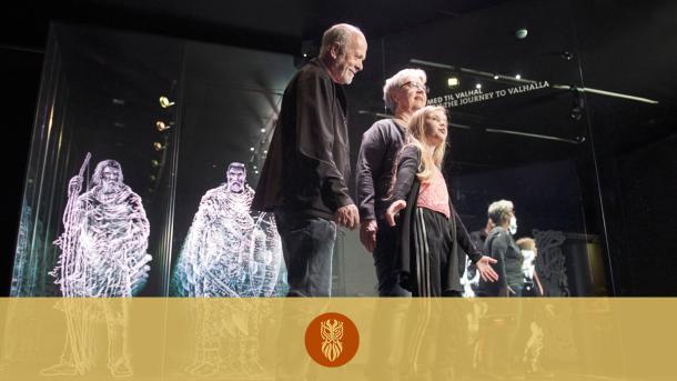 Several older and younger museum visitors form a chain in the interactive exhibition Journey to Valhalla. In the background are glowing Vikings.