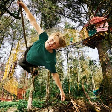 A boy swings from treetop to treetop in the square. There are also long suspension bridges between the tree huts. There is free play here for both adults and children.